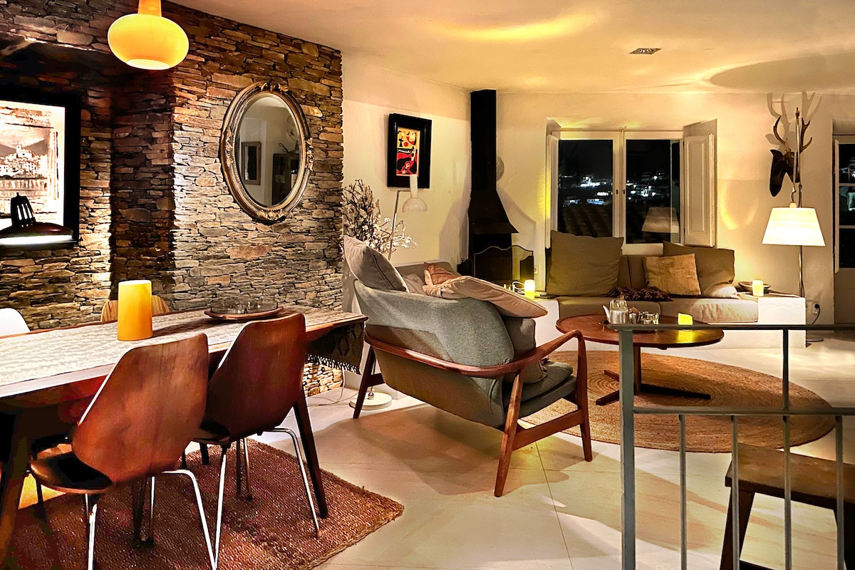 A modern living area features a combination of natural stone walls and soft furnishings. A dining table and stylish chairs are positioned next to a cozy seating area, illuminated by ambient lighting. Large windows allow for natural light and views of the surroundings.