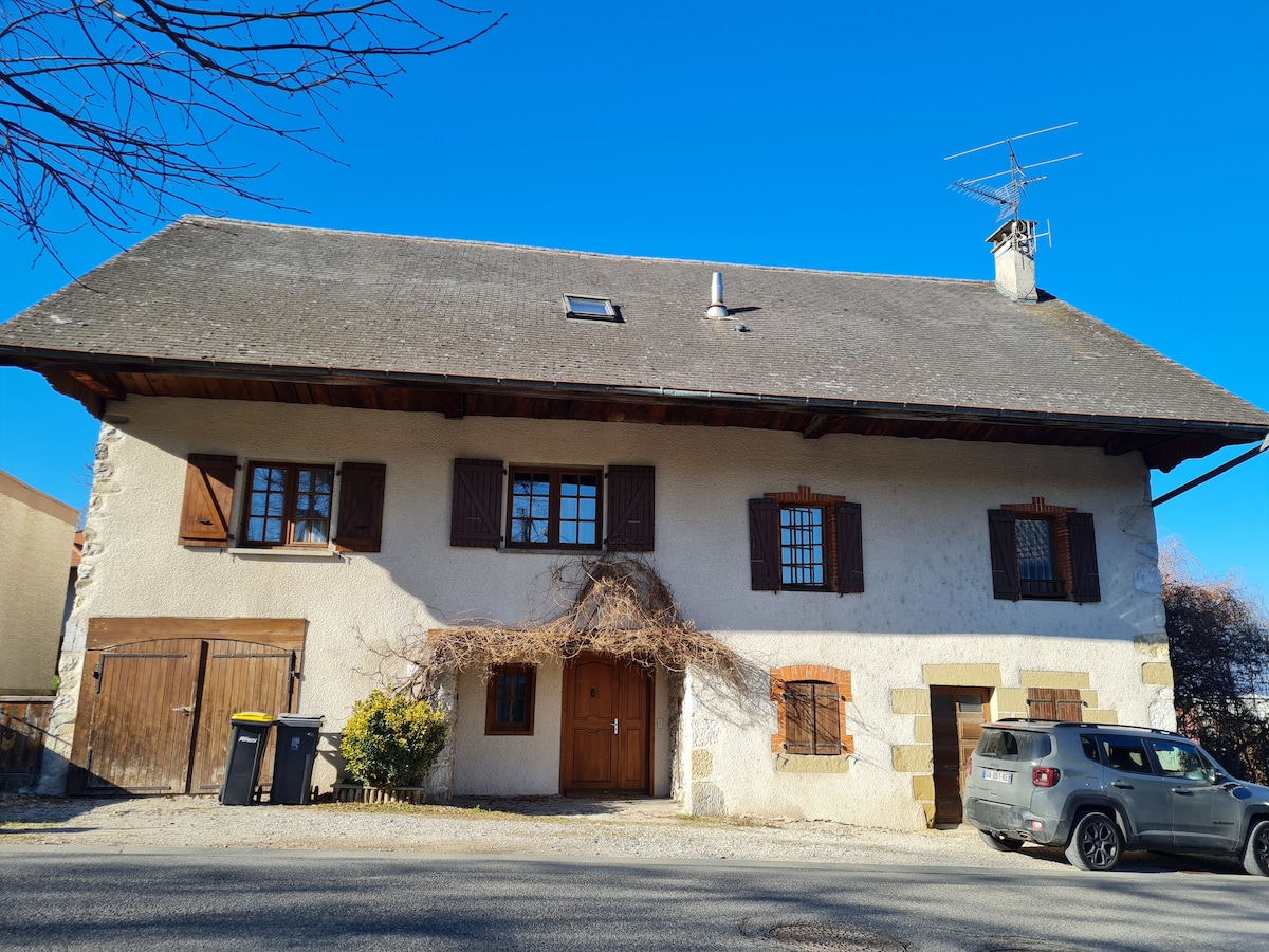 The exterior of a renovated farmhouse displays a combination of traditional and modern elements. The dark wood shutters are contrasted against the light-colored walls. A spacious entrance is framed by wooden doors, with a small garden area visible to the side and a clear blue sky overhead.