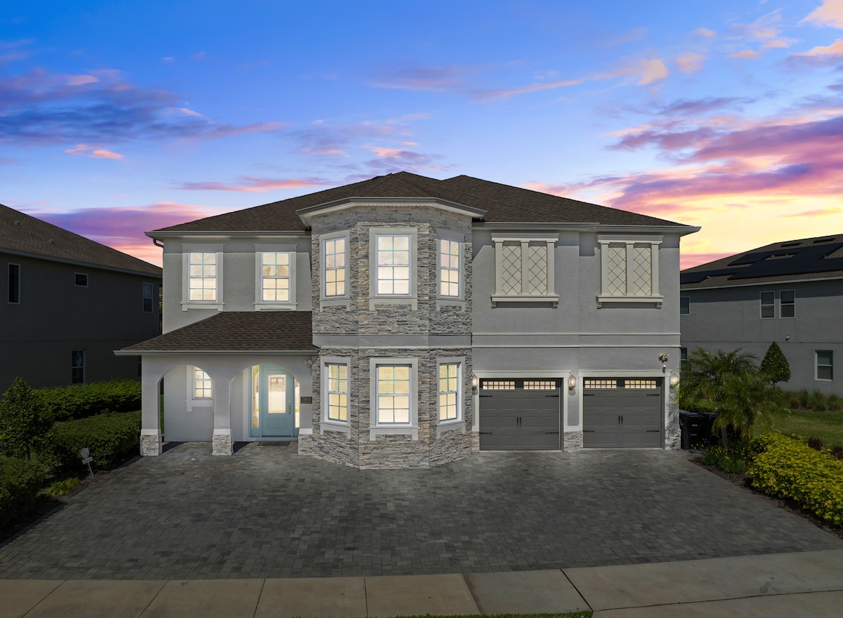 A spacious home is featured at dusk, illuminated by soft lights from its large windows. The exterior blends elegant stone accents with a warm-toned facade, flanked by manicured landscaping. The inviting front porch leads to a double garage, showcasing generous parking space in the driveway.