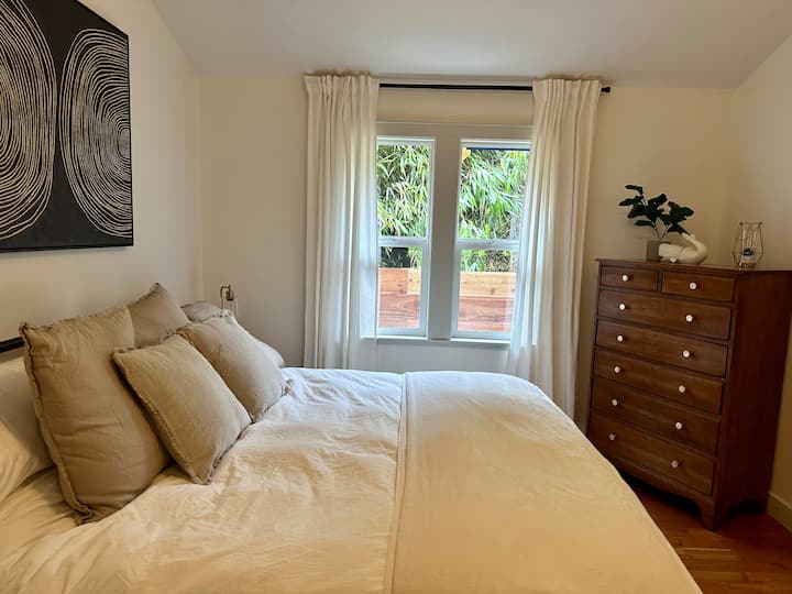 Dormitorio principal con cama tamaño king, lámparas de contacto junto a la cama con puertos USB, cómoda, gran vestidor con puerta de vidrio y espacio de trabajo (silla cómoda, escritorio, lámpara, iluminación y arte de pared) y baño completo con ducha/bañera.