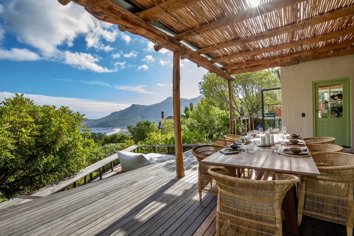 An outdoor dining area features a large wooden table surrounded by woven chairs. The space is covered with a thatched roof, offering shade while allowing views of the surrounding greenery and mountains in the background, with blue skies and a hint of ocean visible.