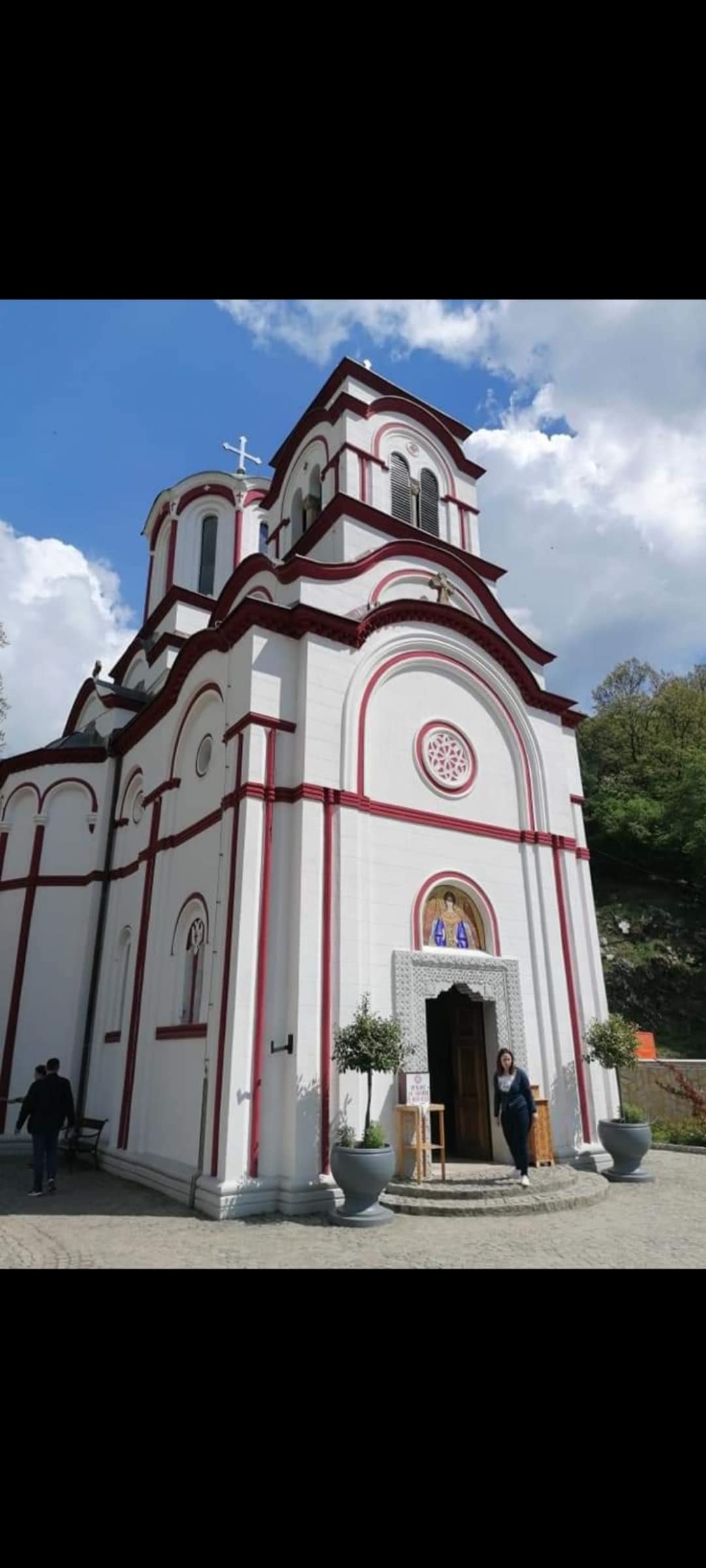 The exterior of a white church is displayed, featuring red accent lines and traditional architectural details. A prominent entrance is visible, framed by planters with greenery. The structure is set against a backdrop of blue skies with scattered clouds.