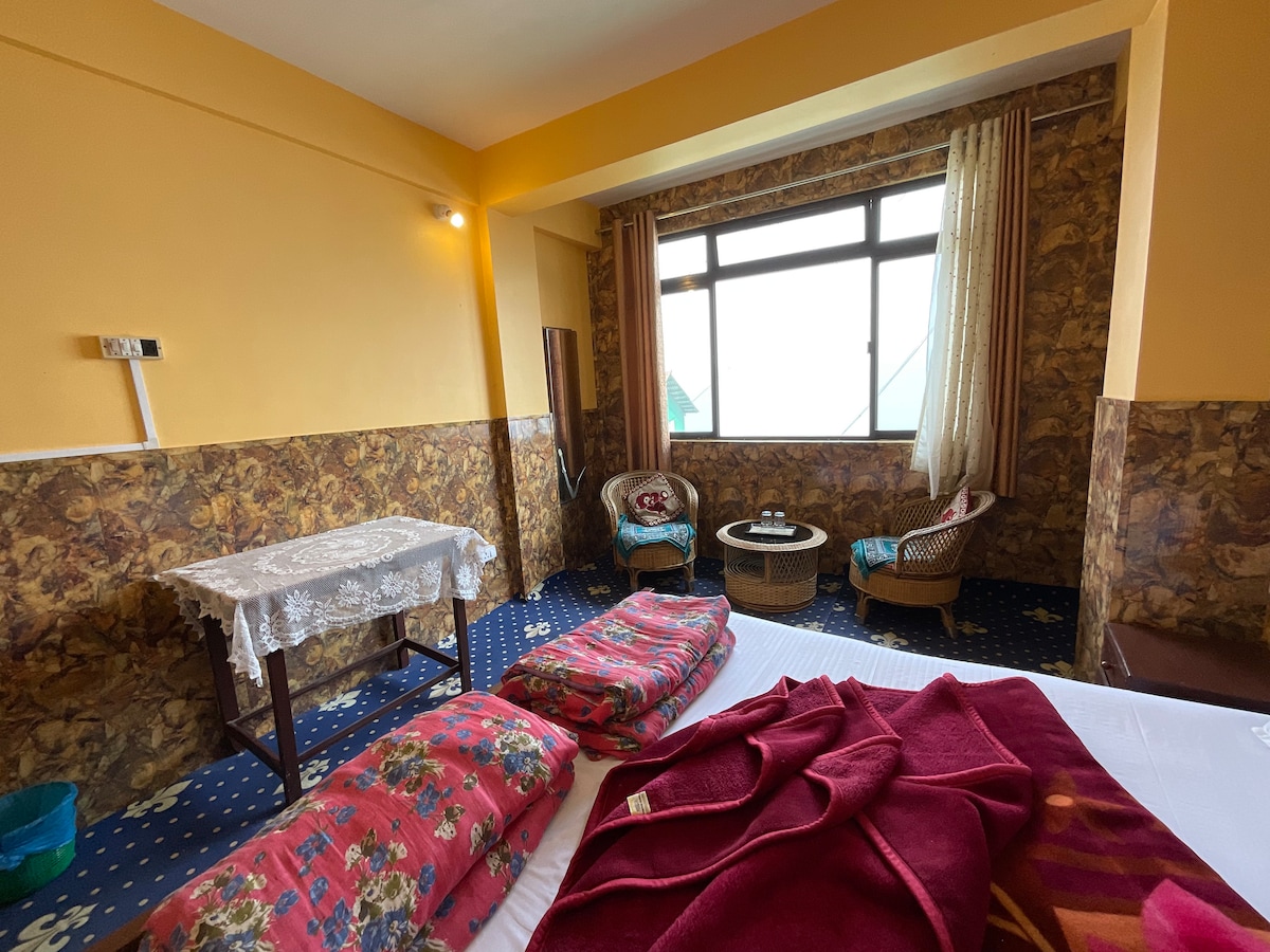 A bright room is presented with warm yellow walls and patterned wallpaper. A window provides a view of the hills, framed by light drapes. Two cozy chairs are positioned near a small round table, while a neatly made bed features blankets and pillows.