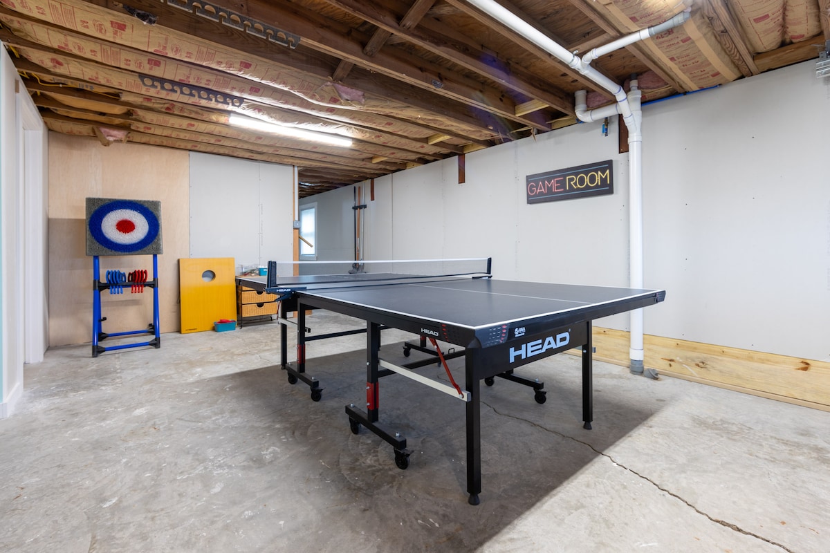 A spacious game room features a championship ping-pong table at its center, surrounded by concrete flooring. A colorful target board and additional games are positioned against the walls, while overhead insulation is visible, contributing to the room’s functional atmosphere.