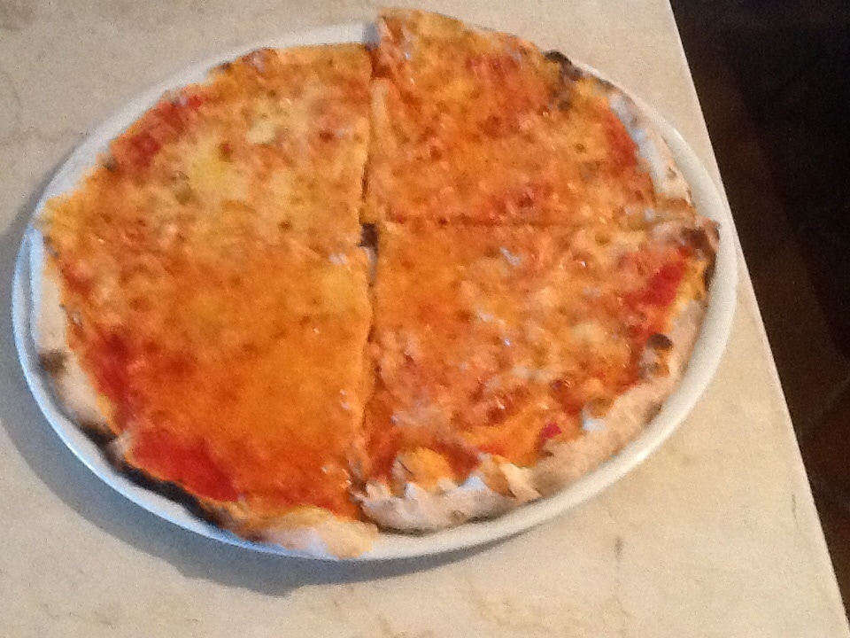 A round pizza is presented on a white plate, featuring a golden-brown crust and evenly melted cheese. The pizza is sliced into four pieces, with tomato sauce visible beneath the cheese. A light marble surface serves as the background.