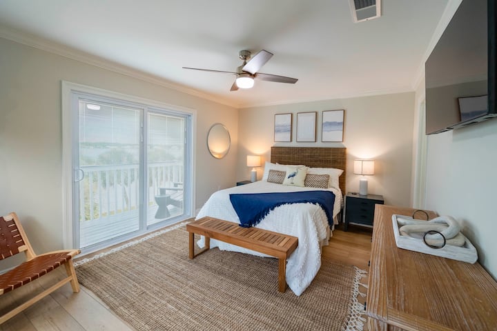 A serene bedroom with coastal decor, featuring a woven headboard, natural wood accents, and sliding doors leading to a cozy balcony view