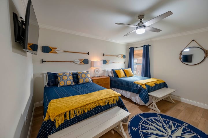A cozy nautical-themed bedroom featuring twin beds with blue and yellow accents, wooden oars as decor, and a compass rug, creating a seaside-inspired atmosphere.