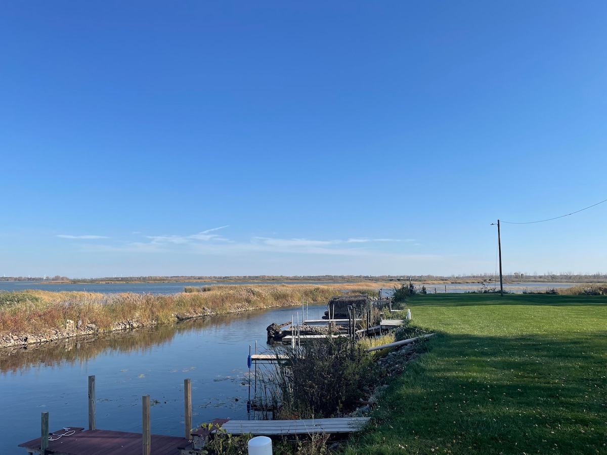 A serene waterside view is presented, featuring two docks by the water's edge. Lush green grass spreads across the foreground, while tall reeds and open water are visible in the background under a clear blue sky.