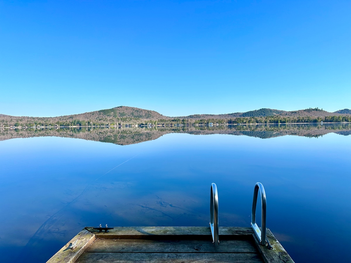 A wooden dock extends over calm waters, reflecting the clear blue sky and surrounding hills. The serene landscape offers a peaceful view, with gentle ripples in the lake providing a contrast to the stillness of the scene.