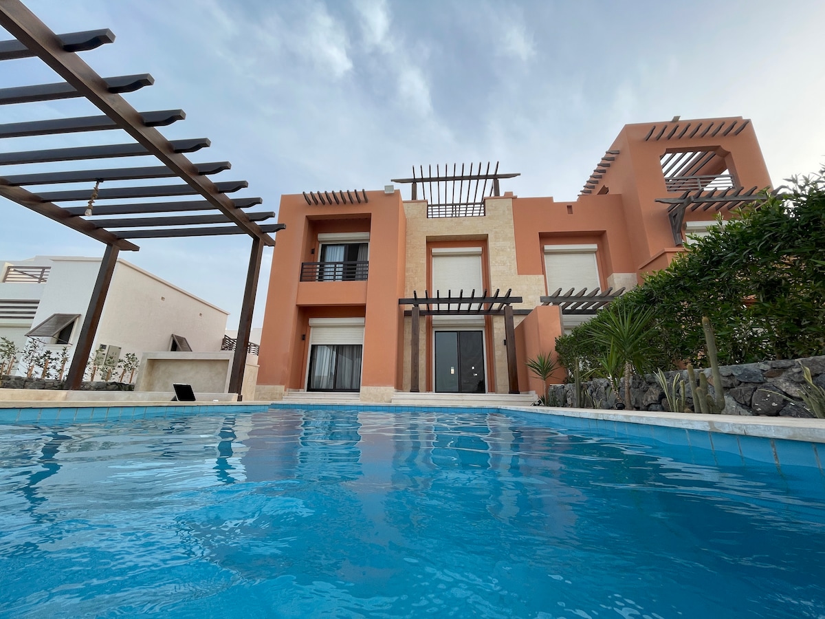The villa's exterior features an elegant design with a terracotta and stone facade. A spacious swimming pool is visible in the foreground, reflecting the surrounding landscape. Wooden pergolas provide shaded areas, enhancing the outdoor living experience.