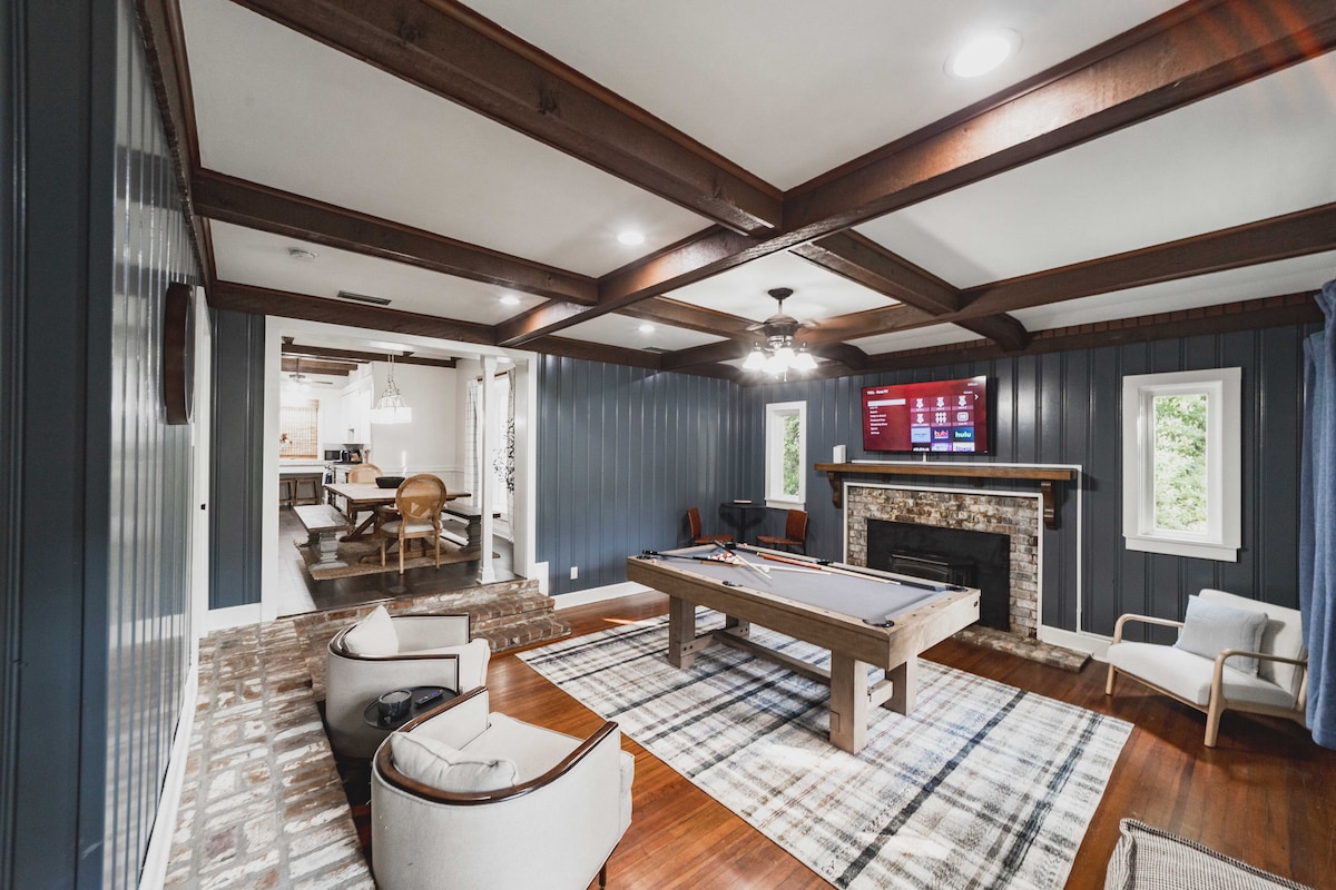 A spacious billiard room features a large pool table centered beneath a decorative ceiling with exposed beams. A cozy seating area with two armchairs and a rug is positioned nearby. Large windows allow natural light, while a television is mounted above a stone fireplace.