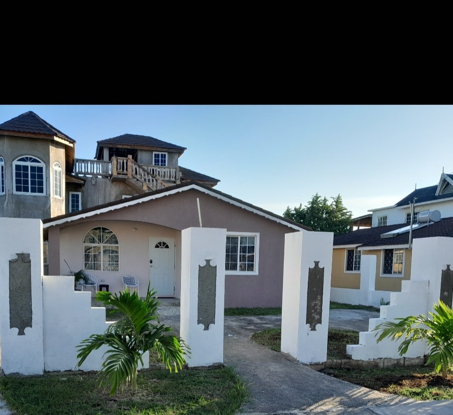 The exterior of a single-story house is visible, featuring a light-colored facade with a welcoming entrance. The yard is bordered by a low wall, with tropical plants adding greenery. Nearby, multi-story buildings are seen, illustrating the community setting.