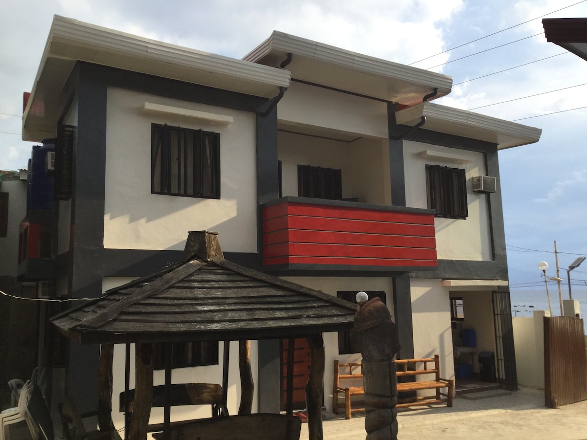 The two-story building features a modern facade with contrasting black and white walls. A striking red balcony extends from the second floor, framed by large windows. A shaded seating area made of wood is positioned in the foreground, enhancing the outdoor space.