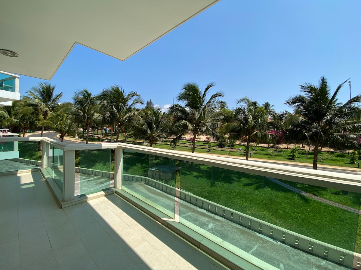 A spacious balcony offers views of lush green lawns and palm trees swaying gently in the breeze. The clear blue sky enhances the tranquil outdoor setting, while the sunlit tile flooring provides a bright and open feel.