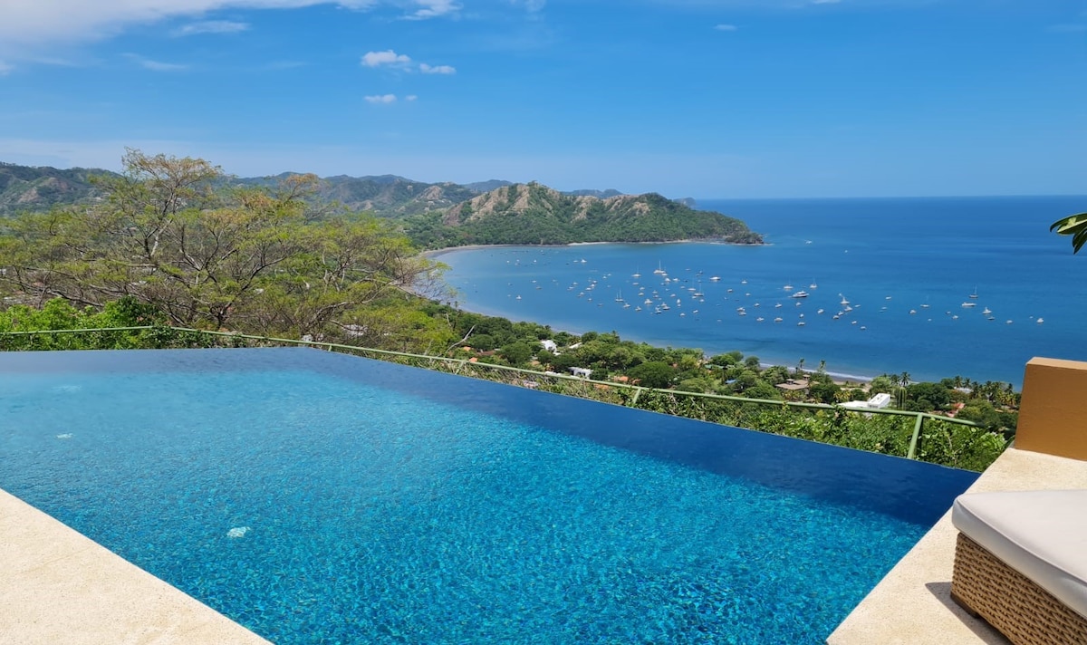 An expansive infinity pool is shown at the edge of a hillside, overlooking a tranquil ocean bay. Clear blue skies and distant green hills frame the serene water, dotted with boats. The pool's reflection mingles with the horizon, creating a seamless view of nature.