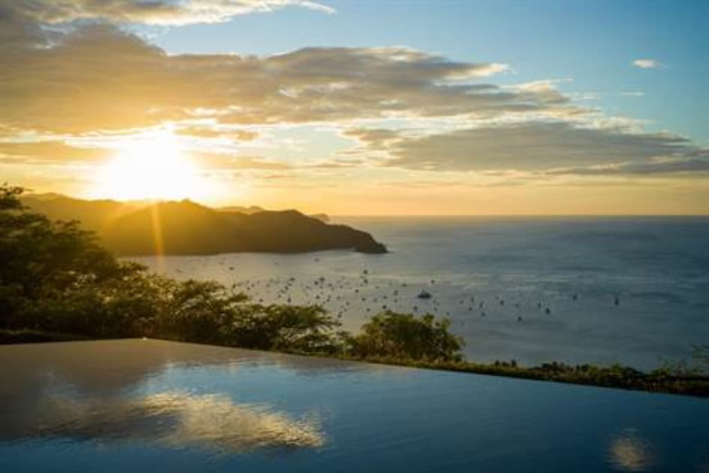 A serene sunset view over the ocean is captured, with gentle waves reflecting golden hues. The infinity pool in the foreground appears to merge with the water, while distant hills outline the coastline, and small boats are visible in the calm sea.