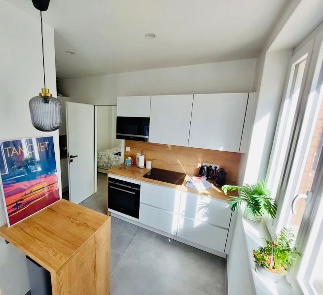 A modern kitchen features sleek white cabinetry and a wooden countertop, complemented by a stylish pendant light. A well-organized layout includes essential appliances, with a subtle green plant adding a touch of nature. Natural light floods the space through large windows.