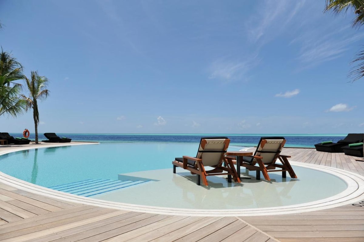 A circular infinity pool is positioned at the edge of the deck, offering unobstructed views of the ocean. Two lounge chairs are placed beside the pool, surrounded by palm trees. A contrasting dark seating area can be seen in the background.