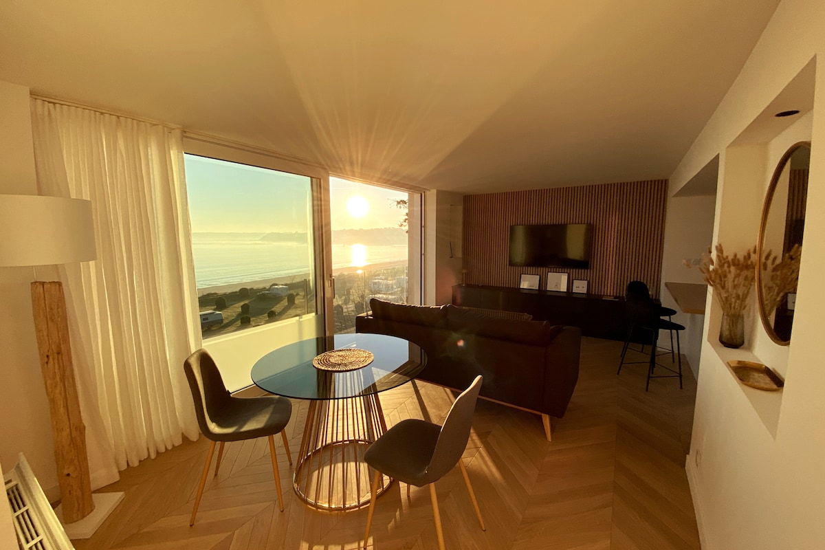 A sunlit living area features a sleek glass table surrounded by stylish chairs. Large glass doors offer a panoramic view of the bay, while a comfortable sofa and a television create a cozy atmosphere. Soft curtains frame the window, enhancing the serene setting.