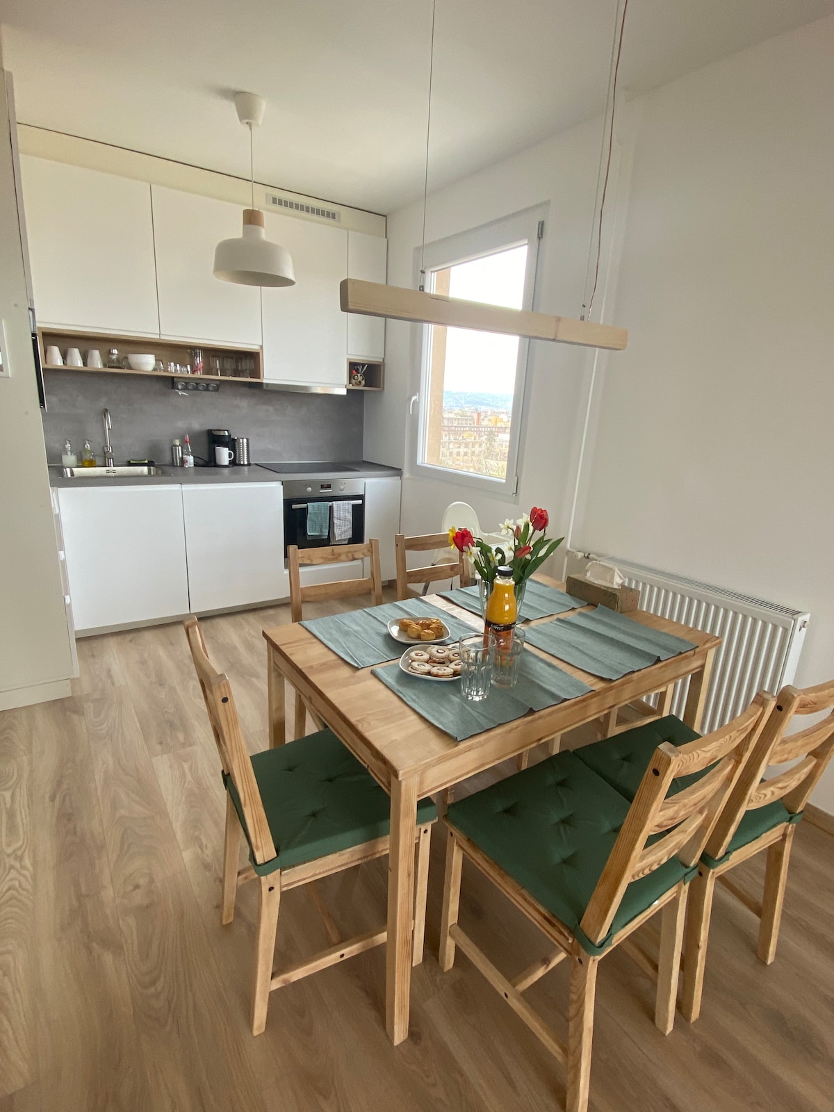 A bright dining area is furnished with a wooden table surrounded by six green cushioned chairs. A small vase of tulips sits at the center alongside a plate of cookies. The kitchen is visible in the background, featuring modern cabinetry and a view from the window.