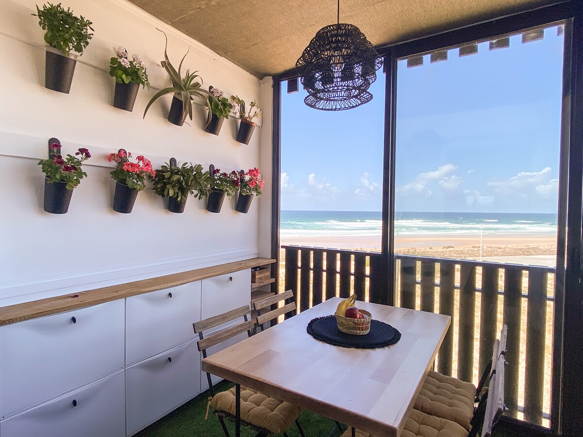 A bright dining area features a table surrounded by chairs, positioned near a large window offering ocean views. Decorative plants are arranged on the wall above a countertop, enhancing the space with greenery. Natural light fills the room, creating an open and airy atmosphere.