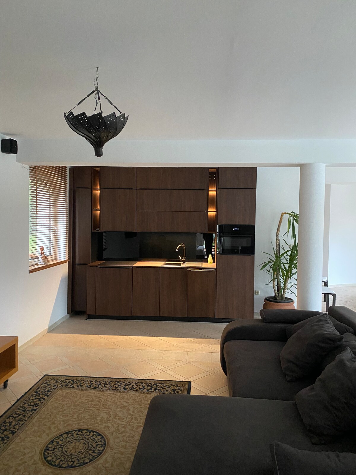 A modern kitchen is featured with sleek wooden cabinetry and integrated appliances. Soft lighting highlights the countertops. A comfortable seating area with a dark sofa is visible in the foreground, complemented by a patterned rug on the tiled floor and a potted plant nearby.