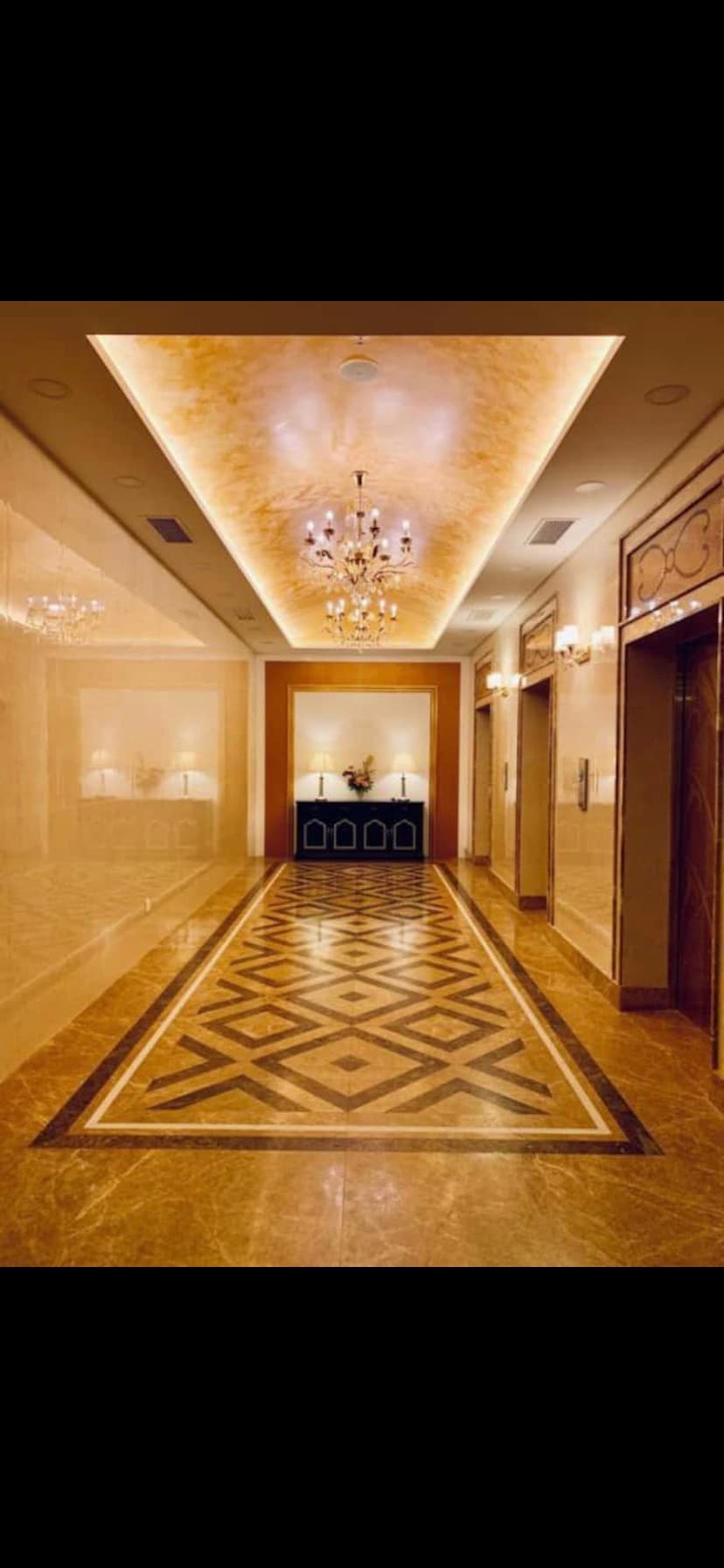 A modern hallway features elegant lighting from a chandelier above, with polished marble floors displaying geometric patterns. Decorative wall panels and soft lighting create a refined atmosphere, while elevator doors are present along one side.