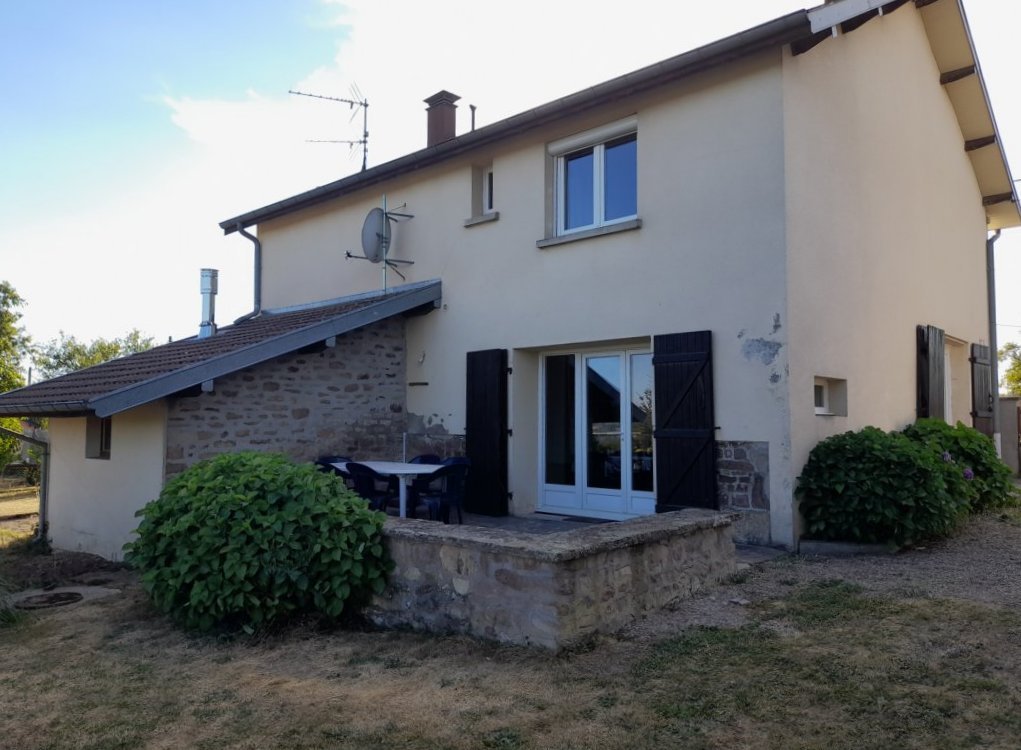 A two-story house is highlighted, constructed with a combination of stone and smooth exterior walls. Outdoor seating is available on a small terrace, surrounded by greenery and gravel. The house features large windows and shutters, providing a blend of rustic and modern elements.