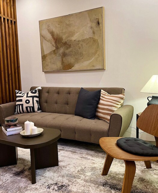 A cozy living area is showcased with a beige sofa adorned with decorative cushions in various patterns. A round coffee table holds books and a trio of candles. A modern accent chair complements the soft textures, while a piece of abstract wall art adds visual interest to the space.