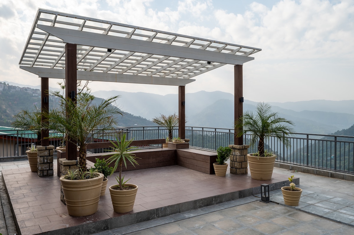 A rooftop terrace features a pergola providing shade, surrounded by potted plants. The area offers panoramic views of distant mountains and valleys, creating a serene environment for relaxation and gathering.