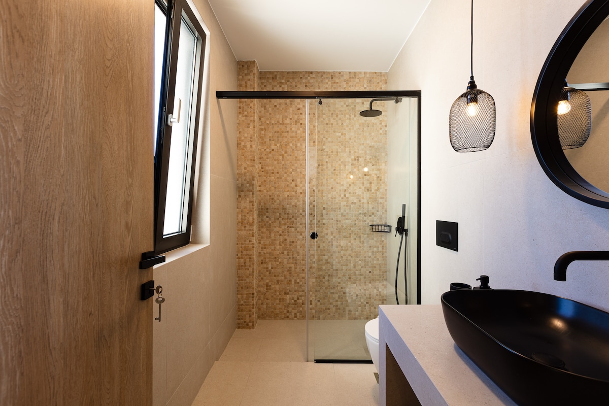 A modern bathroom features a glass shower enclosed by textured tiles. Natural light enters through a window, illuminating the space. A sleek black sink is complemented by a dark wall mirror and an elegantly designed light fixture, enhancing the contemporary design.