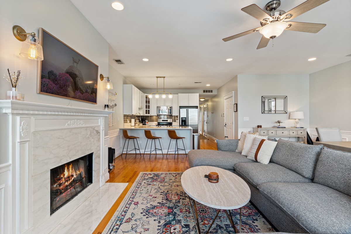 A spacious living area features a cozy fireplace set into a white marble surround. Light wooden floors and neutral walls create a calming backdrop. A large sectional sofa faces a coffee table, while stylish bar stools complement the nearby kitchen island.