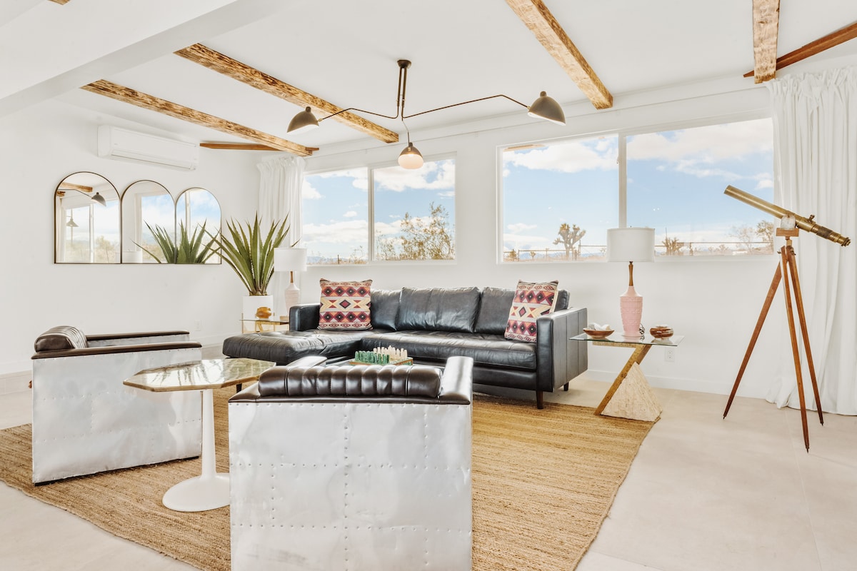 A bright living space features large windows offering views of the desert landscape. A sleek, dark leather sofa faces two contrasting lounge chairs, complemented by a round side table. Decorative pillows and a telescope enhance the inviting ambiance, with light streaming through sheer curtains.