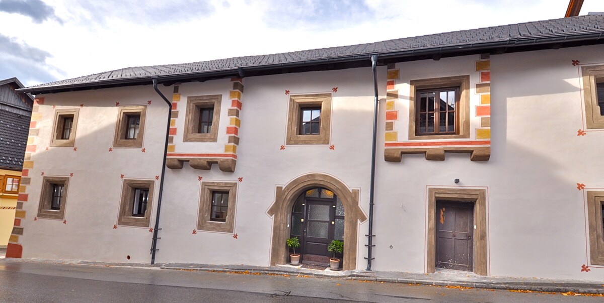 The exterior of the historic house is showcased, featuring a smooth plaster façade with decorative patterns. Windows with wooden frames are present on both levels. The entrance is highlighted by an arched doorway flanked by symmetrical potted plants, reflecting the building's traditional architectural style.