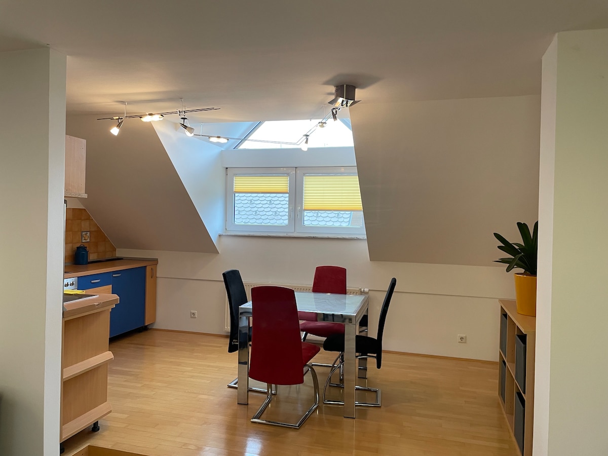 A modern dining area features a glass table surrounded by six colorful chairs. Natural light enters through two skylights and a window adorned with yellow blinds. A wooden kitchen counter is visible in the background, complemented by blue cabinetry.