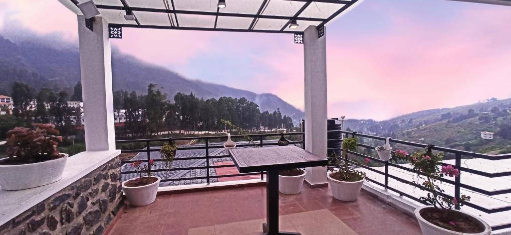 An outdoor dining area is featured under a large, shaded terrace. A wooden table is positioned centrally, surrounded by potted plants. The mountain landscape and soft pastel sky are visible in the background, creating a serene setting for meals or relaxation.