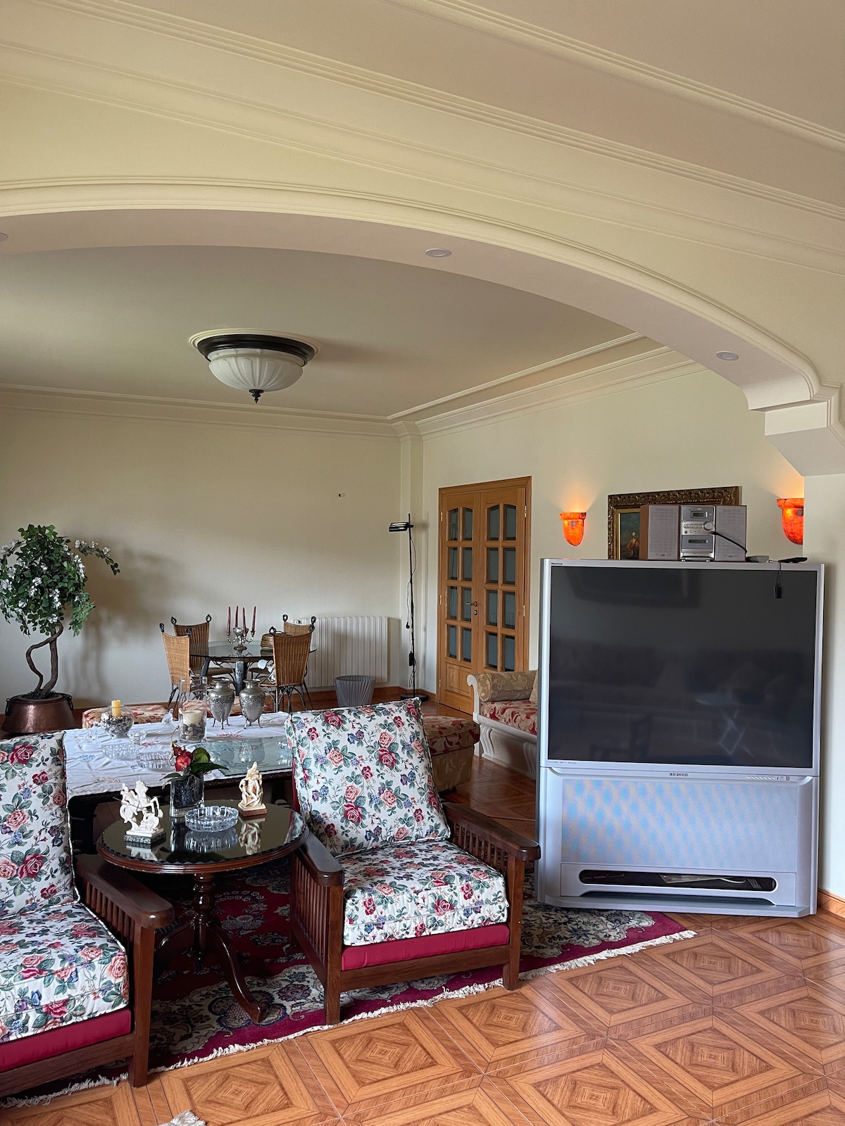 The living area features vintage-style furniture with floral upholstery, including two plush armchairs and an elegant coffee table. A large television is positioned beside a decorative cabinet, and a dining table with chairs is visible in the background, creating a welcoming atmosphere.