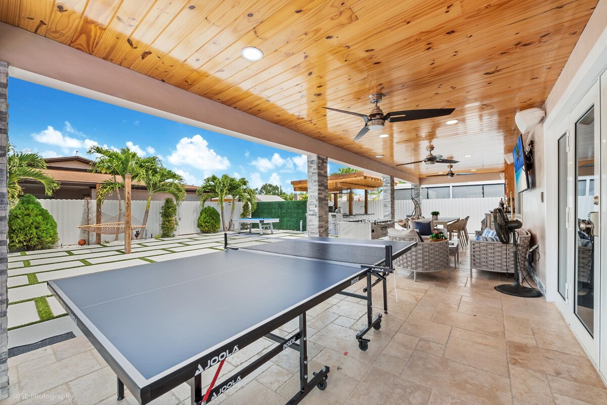 An outdoor entertainment area features a ping pong table positioned under a wooden ceiling with ceiling fans. Surrounding this space are well-maintained pavers and manicured landscaping, creating a pleasant environment for leisure and relaxation.