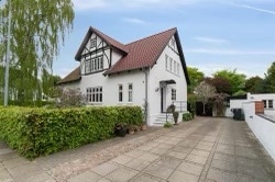 A charming two-story house features a combination of white and dark timber accents. The inviting entrance is flanked by well-maintained greenery, and a paved driveway leads to the front door. The serene surroundings reflect a peaceful neighborhood setting.
