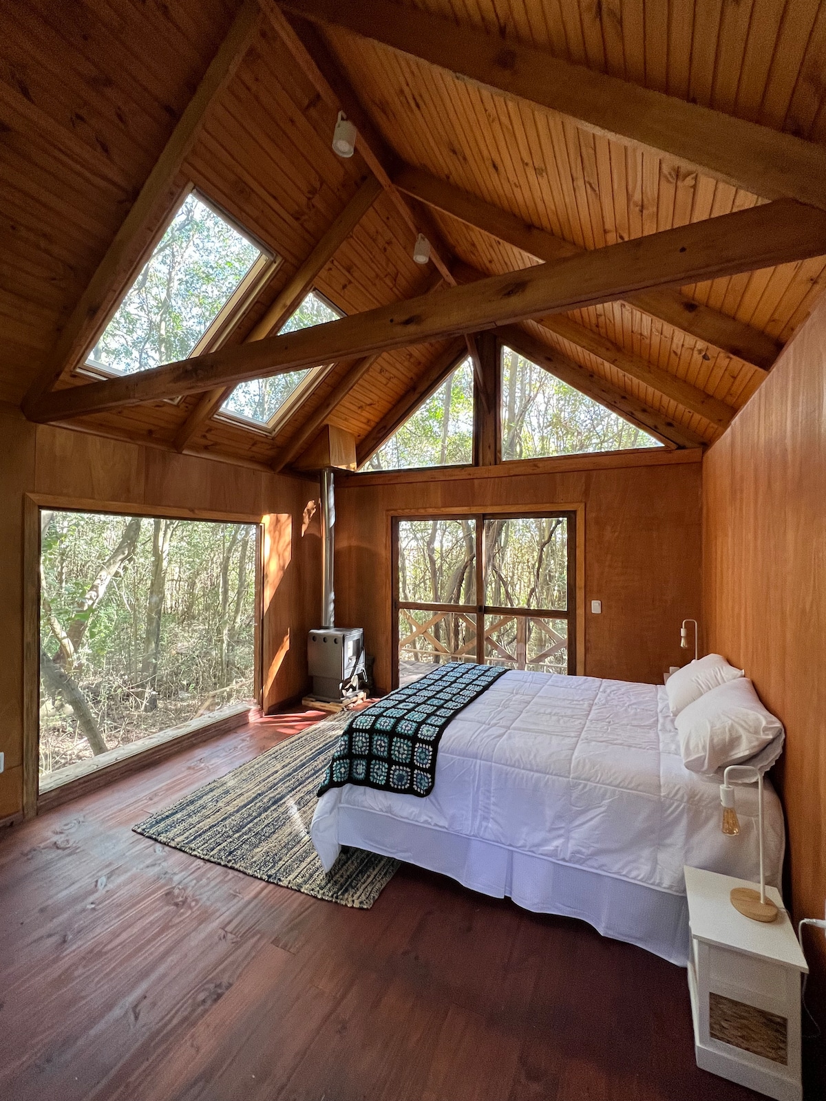 A cozy cabin interior features a spacious bed dressed in white linens, accented by a textured black and white blanket. Large windows invite natural light and views of the surrounding trees, while wooden beams highlight the ceiling's rustic charm.
