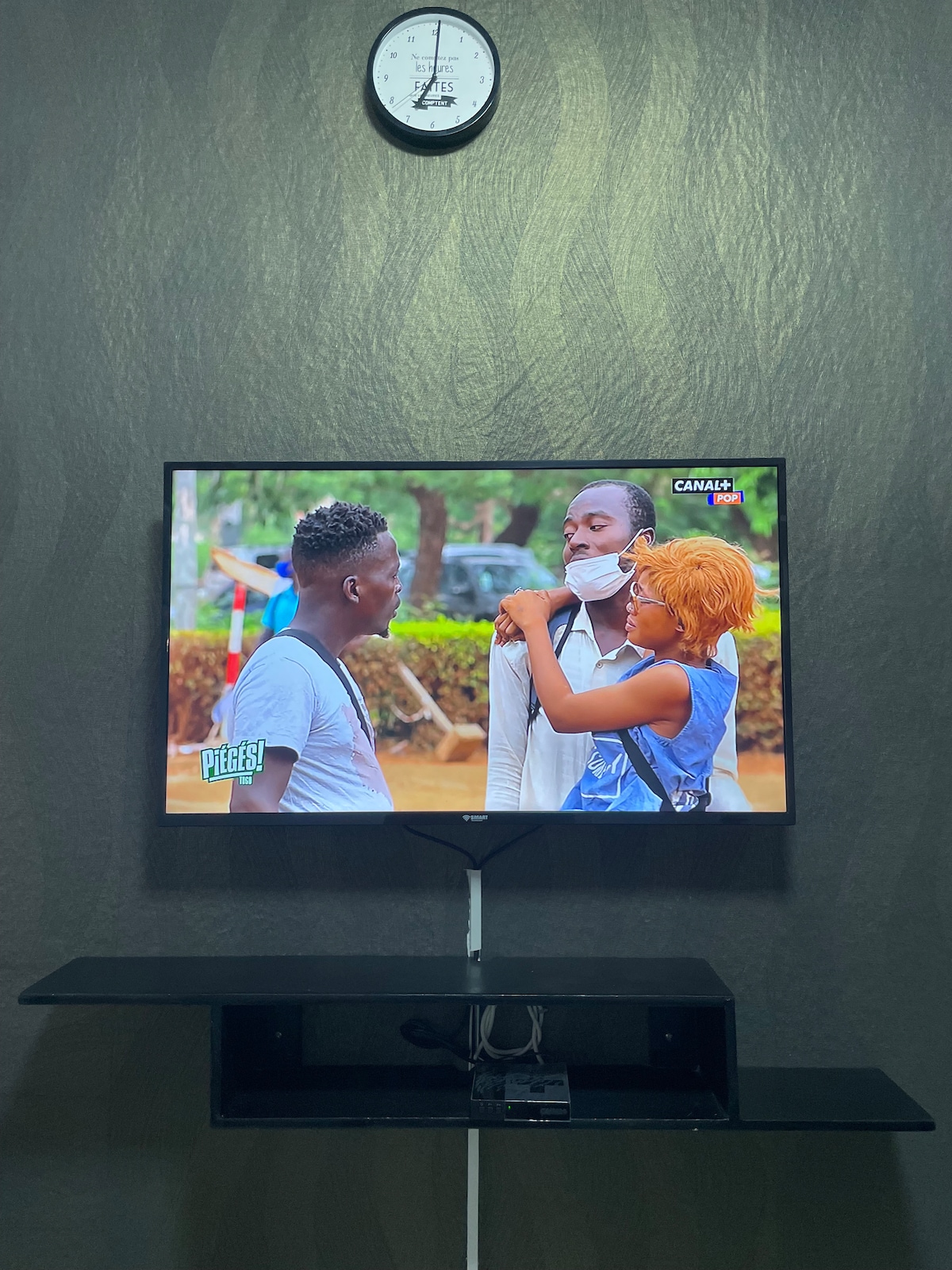 A television is mounted on a dark wall. Below it, a sleek, minimalist TV stand holds the device. A clock is placed above the television, indicating the time. The image on the screen shows two individuals engaging in conversation in an outdoor setting.