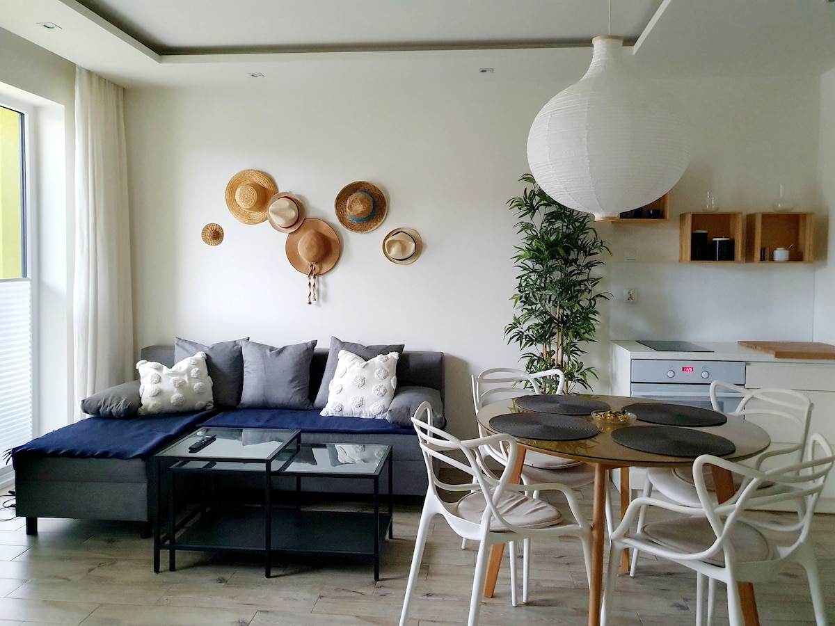 A modern living area features a comfortable corner sofa adorned with decorative pillows, paired with a coffee table. A dining table set for four is visible nearby, complemented by stylish white chairs. Wall-mounted hats add a decorative touch, with greenery enhancing the ambiance.