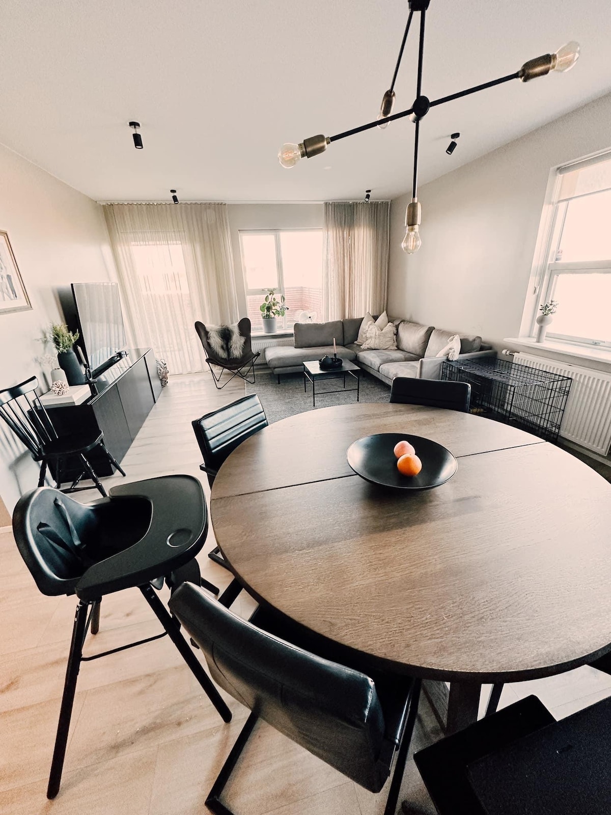 A dining area features a round wooden table surrounded by chairs, with a bowl of oranges as the centerpiece. A cozy living space is visible in the background, showcasing a comfortable couch, modern lighting, and large windows with soft curtains that allow ample natural light.