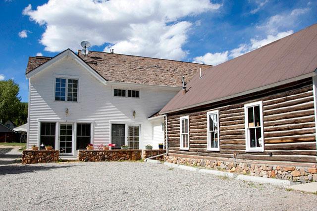 The exterior of the lodge combines modern and rustic elements, featuring a two-story white structure alongside a traditional log cabin. A gravel area is present in front, with seating visible near large windows that invite natural light.
