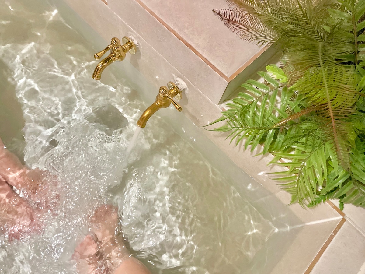 A jacuzzi is filled with clear water, showcasing its smooth, tiled edges. Two elegant golden faucets are seen at the top, alongside lush green ferns that add a touch of nature to the serene setting.