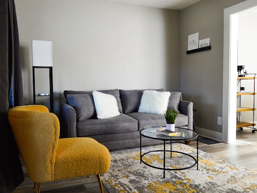 A cozy living area features a gray sofa adorned with white cushions, complemented by a yellow accent chair. A round coffee table, placed on a textured area rug, is topped with a small plant. A door leads to another room in the background.