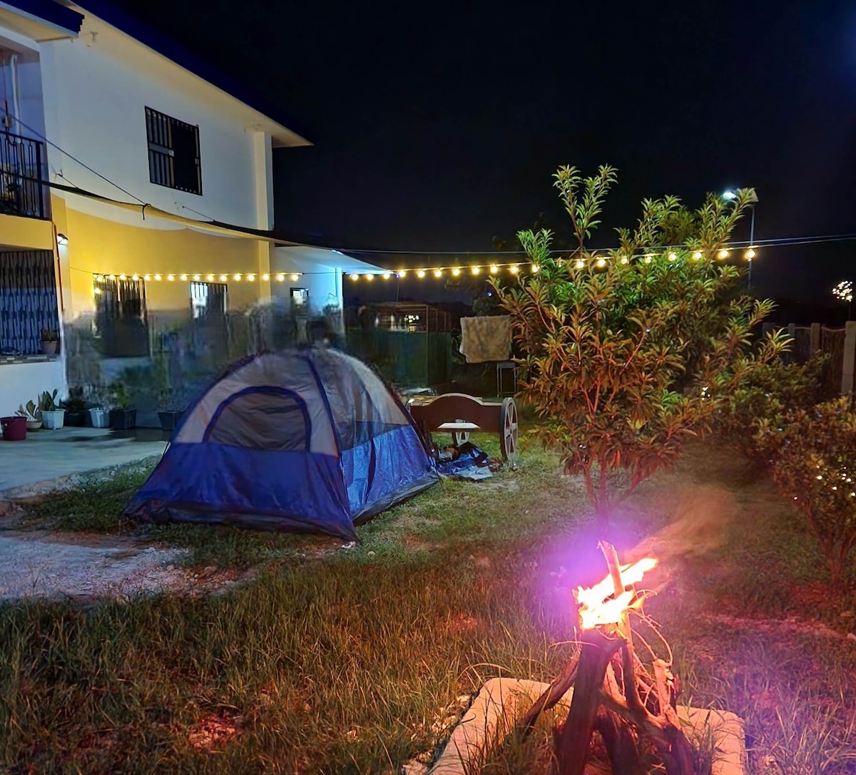 A camping area is illuminated by string lights, featuring a blue tent pitched on grass beside a small campfire. Nearby, a tree provides natural elements to the outdoor space, while the building stands in the background, creating a cozy atmosphere for outdoor activities.
