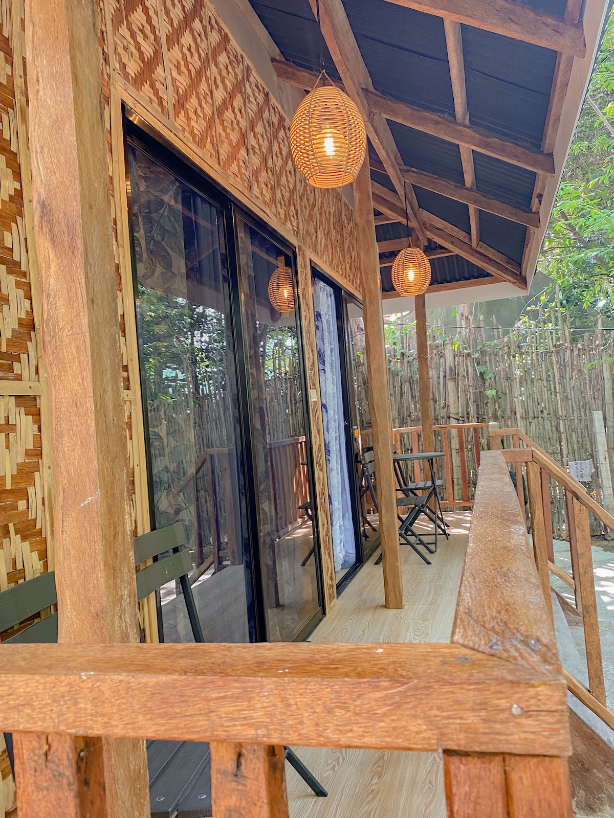An inviting outdoor balcony area features natural wood railings and floorboards. Two black folding chairs are positioned to enjoy the surrounding greenery. Soft lighting fixtures hang from the roof, adding a cozy touch to the space. Lush bamboo is visible in the background.