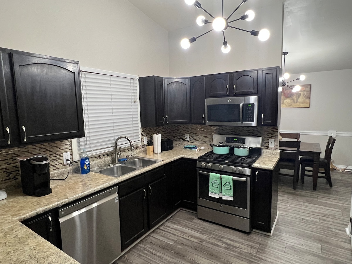A modern kitchen features dark cabinetry and a spacious countertop. Stainless steel appliances are present, including a gas stove and a refrigerator. A stainless steel sink is situated below a window, while a dining area with wooden chairs can be seen in the background.