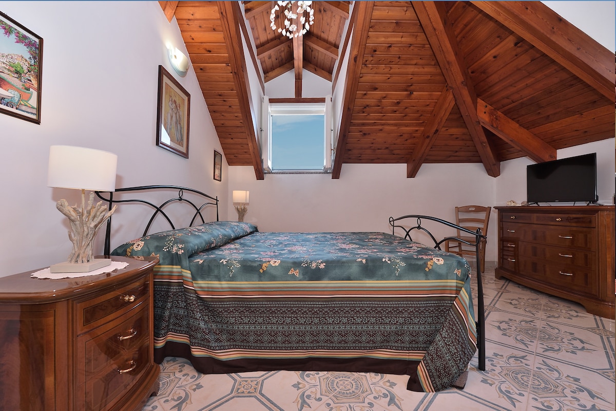 A spacious bedroom features a patterned bedspread and is illuminated by natural light coming from a window above. Wooden beams create a warm atmosphere. An elegant lamp and a wooden nightstand are present beside the bed, while a dresser and a television complete the room's furnishings.